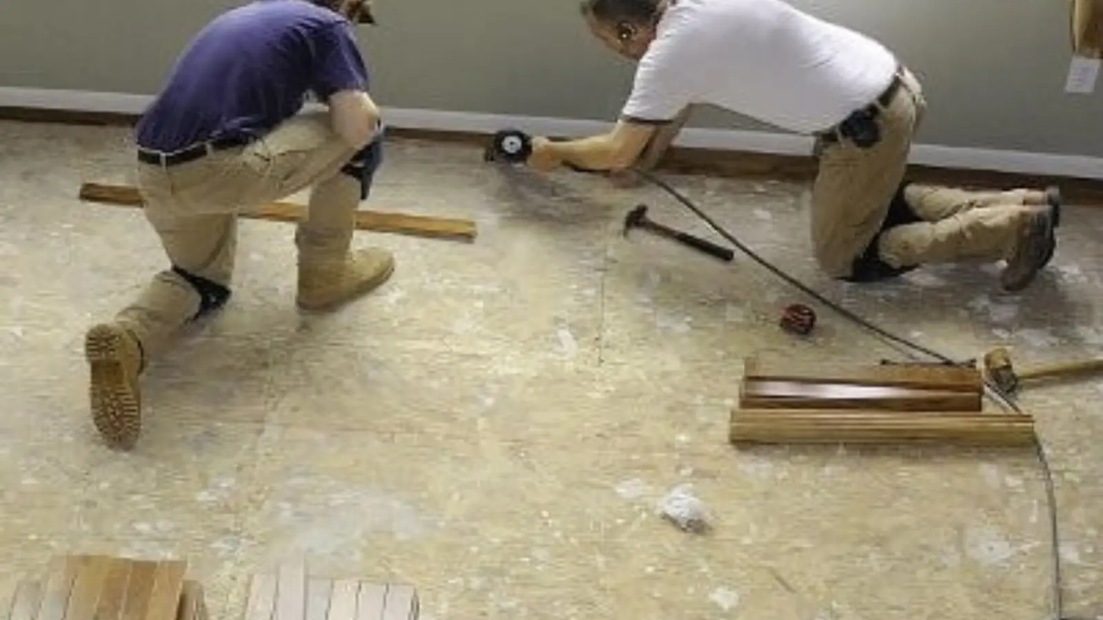 Two workers kneeling on a stripped subfloor installing replacement flooring boards